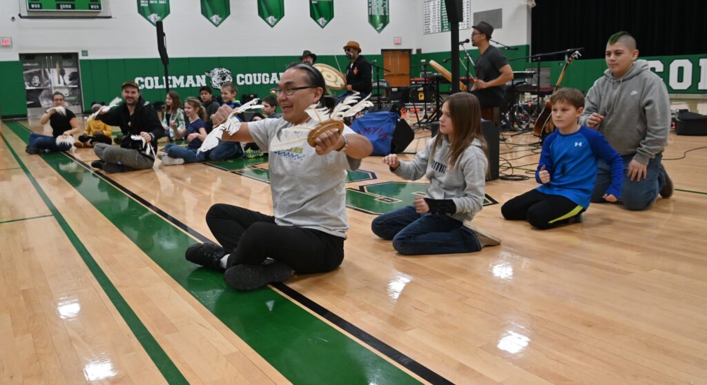 Adults and children sit in imaginary kayaks on the floor of an elementary school gym.