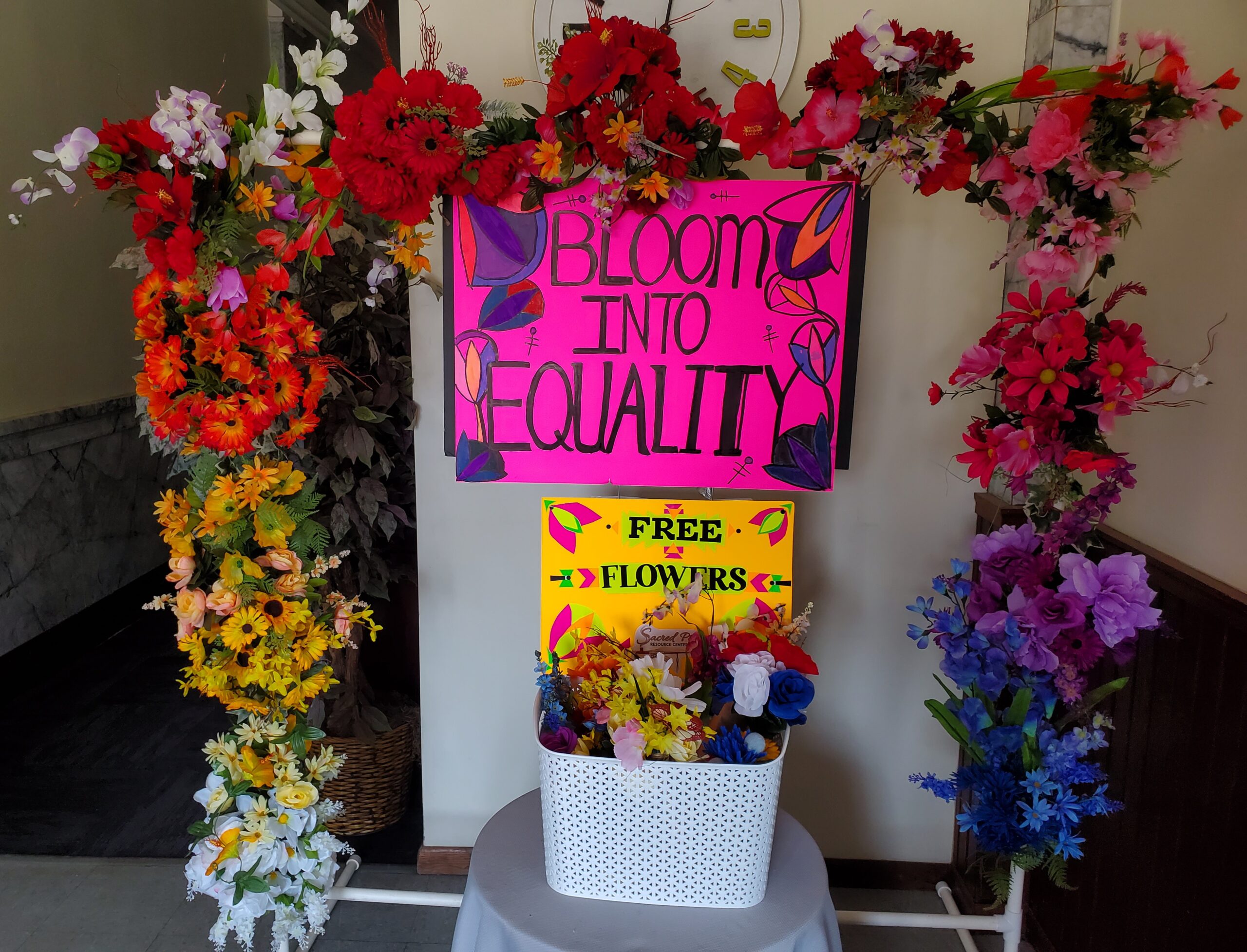 A colorful flower arch that surrounds a sign that says Bloom into Equality and a bucket offering free flowers