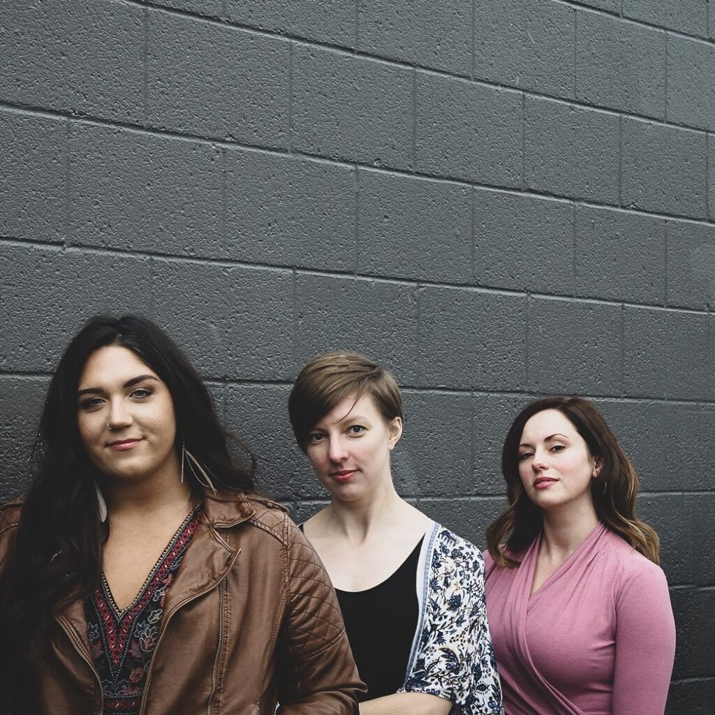 Good Time Girls, a folk trio from Ohio, pose together against a brick wall with soft smiles on their faces.