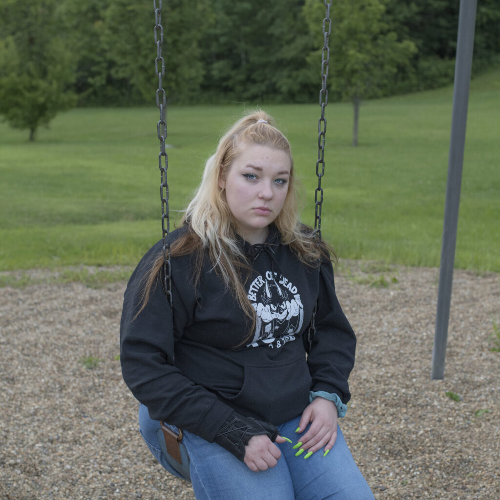 A young person wearing a sweatshirt and jeans, sits on a swing and looks at the camera with a serious expression.