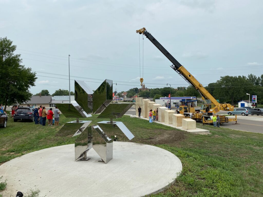 A reflective 8 point star sculpture stands on a circular cement slab in the middle of the grass. In the background, there's a bulldozer and people surrounding another sculpture in progress.