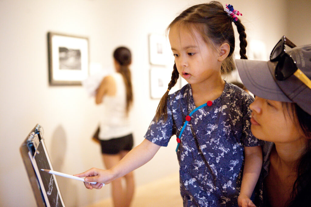 In an art gallery, a person crouches behind a child who is using a white color pencil to write on a black piece of paper.
