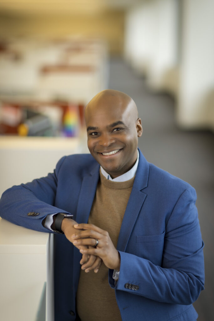 Headshot of a smiling person of medium dark skin tone, who is bald and wearing a brown sweater under a blue suit jacket, with hands crossed and leaning against a cubicle.
