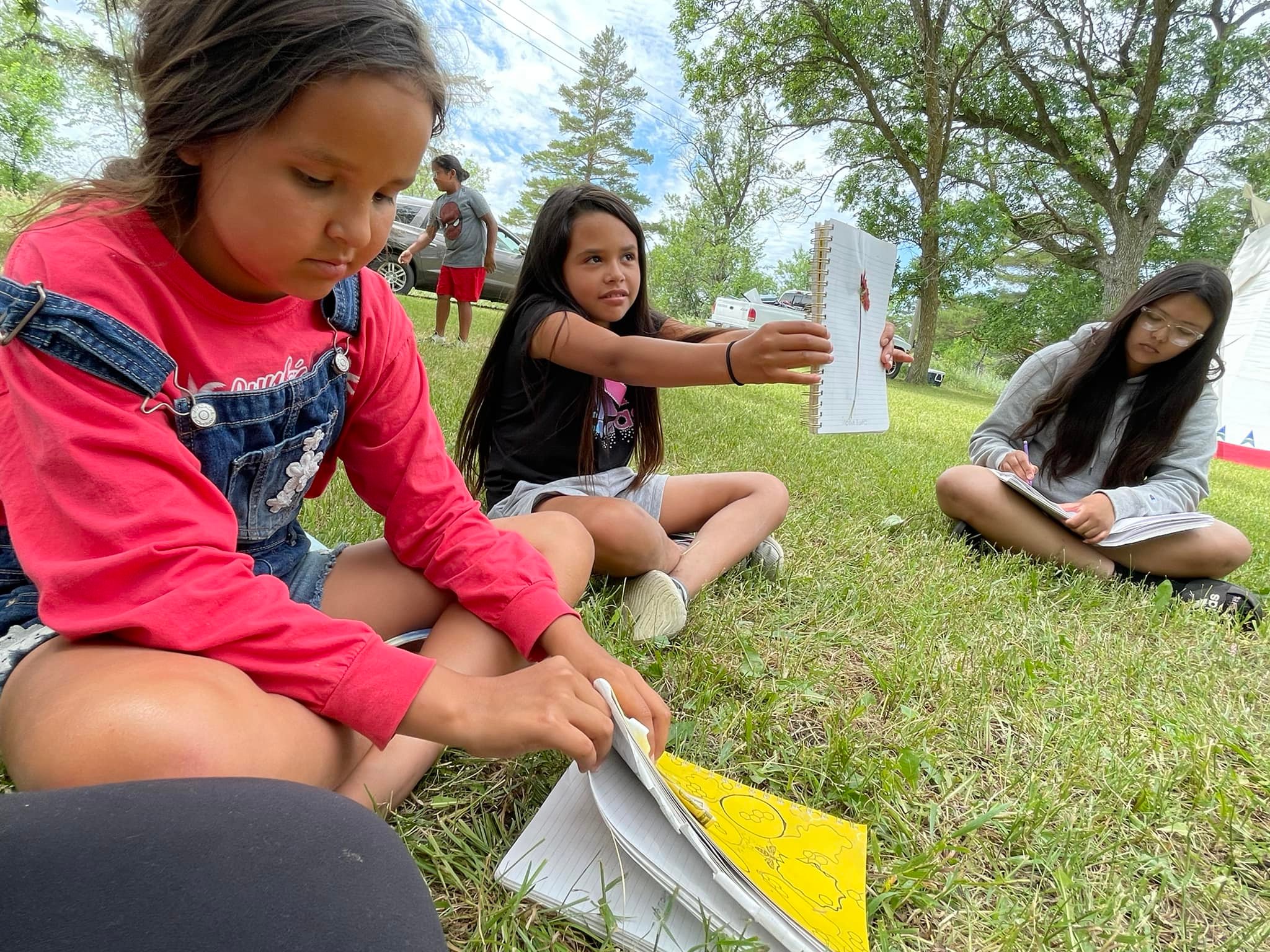 Three children sit in a line on the grass with notebooks. The child in the center holds her notebook up to show a wildflower that she's stuck to the page.