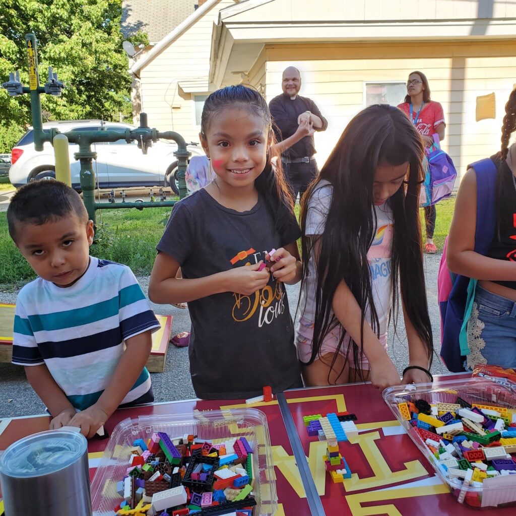 A few children are standing at a table with two containers of Legos, and the one in the middle is smiling at the camera.