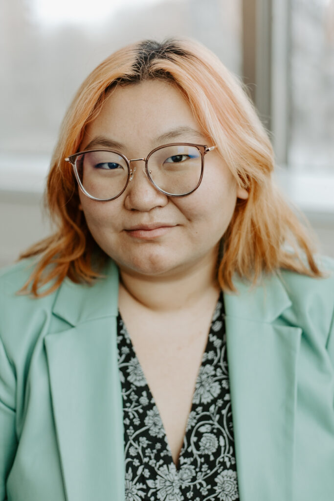 A smiling person of medium light skin tone, with light brown rimmed glasses and shoulder length pink hair, wearing a seafoam green blazer over a black and seafoam green patterned top