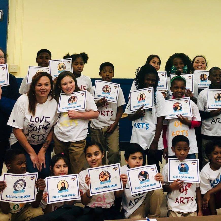 Students and a teacher are gathered and posing for the camera, all wearing t-shirts that say "Love of the Arts" and holding up participation award certificates.