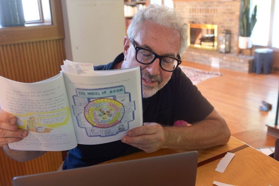 A person sits at a table in front of a laptop, smiling and holding up a book open to a page that reads "Wheel of Doom" at the top.