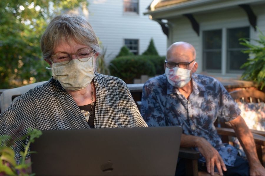 Two older people wearing protective cloth masks sit on a sunny patio, smiling and looking at a laptop.