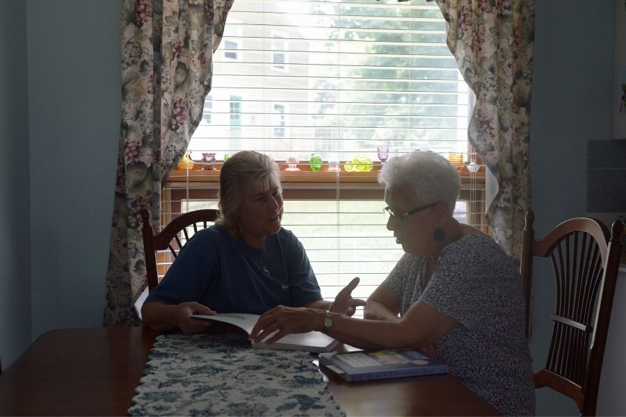 A mother and daughter sit at a kitchen table together, cast in the light from the kitchen window. They're looking at a book together and talking.