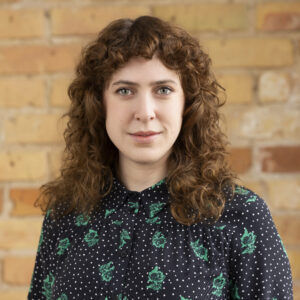 Headshot of a smiling person of light skin tone, with light brown curly hair, and wearing a patterned black and green top