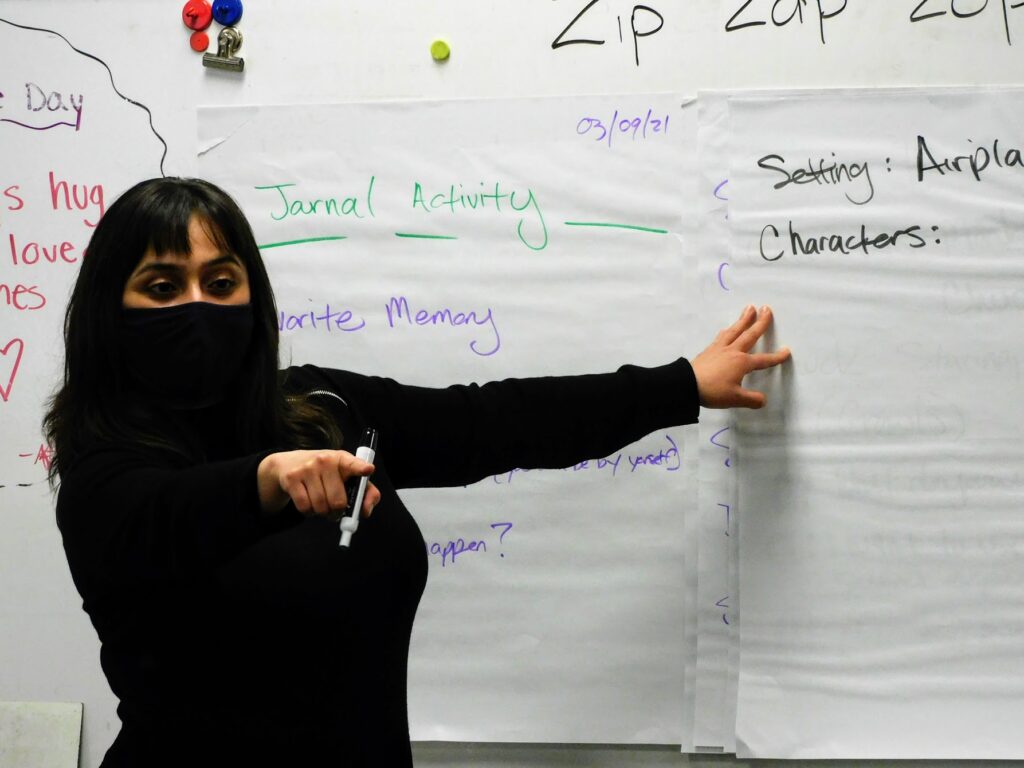 A person of medium skin tone and long black hair, and wearing a black cloth mask and shirt, is pointing at something off camera with a marker in hand, with the other hand touching poster paper.