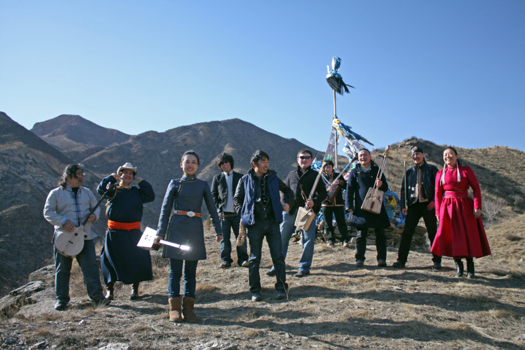 Anda Union, a band based in China who primarily specialize in khoomii (a traditional type of Mongolian overtone singing), pose in the middle of a mountain range, holding various instruments.