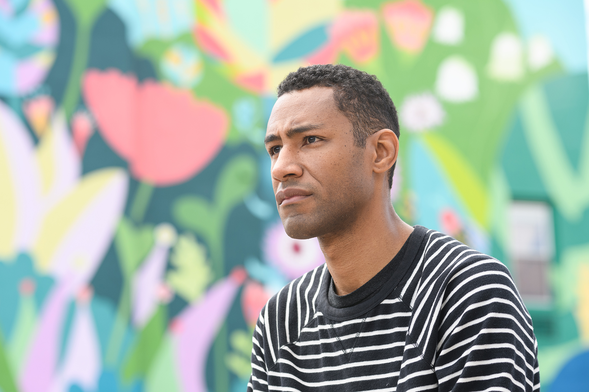 Headshot of a person of medium dark skin tone with short curly black hair, wearing a black and white striped shirt, and sitting in front of a vibrant flower mural.