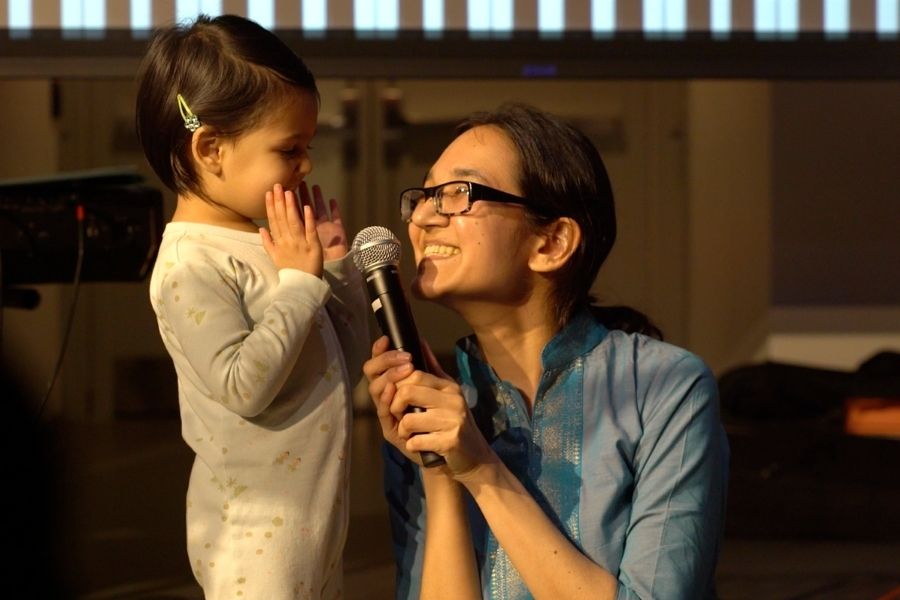 An adult with a big smile kneels on the ground, and is holding a microphone to a small child’s face. The child has her hands up on either side of their mouth to speak into the microphone.