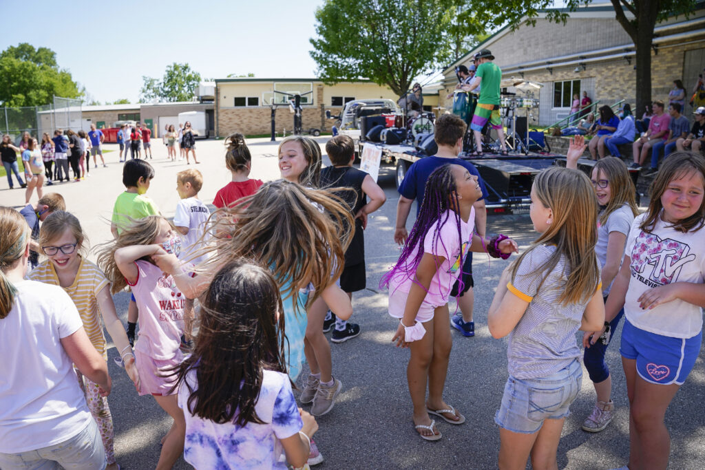 Children gather and dance around in a parking lot while a band performs on a stage behind them.