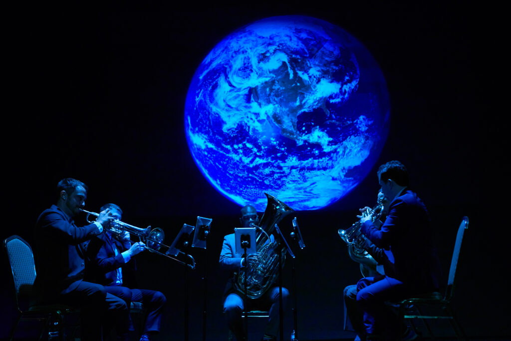 Axiom Brass sits on a stage and play various brass instruments, below a glowing projection of planet Earth.