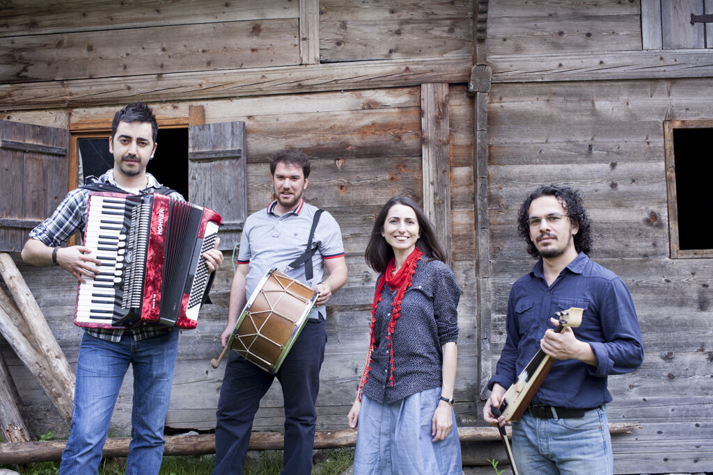 Aysenur Kolivar, band from Turkey who are heavily influenced by Anatolian Black Sea culture, pose outside of a wooden house. They hold various musical instruments including an accordion and a drum.