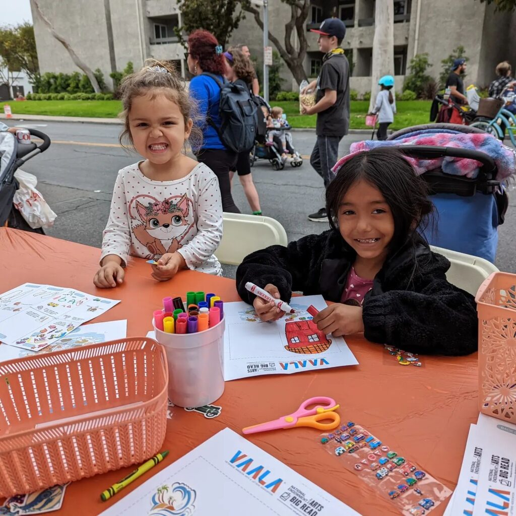 Two smiling children drawing and coloring at a table as people walk down the street behind them.