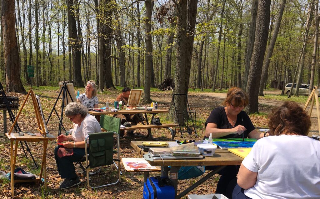 People sit at tables outside in the woods, painting on canvases.
