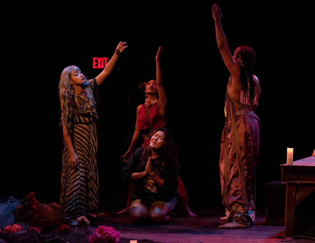 Four people perform on a dimly lit stage; three of them stand looking up with one arm stretched toward the sky, and one sits in the center with hands up in prayer.