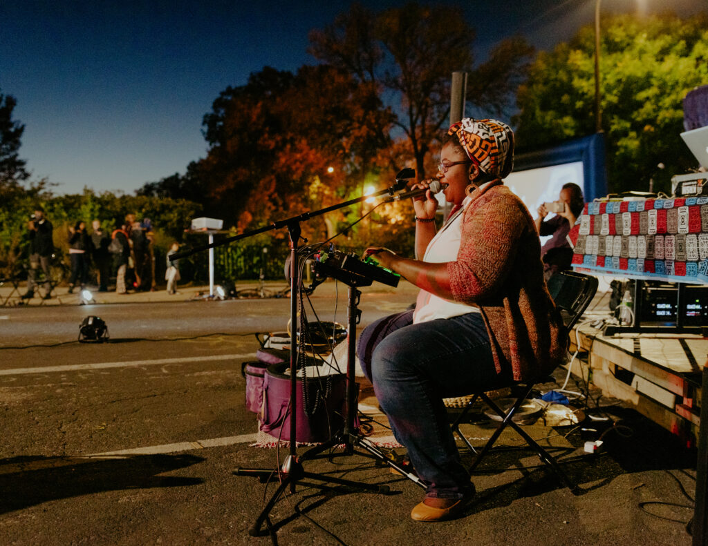 A person performs on the side of a road at dusk, with a keyboard in front of them and singing into a microphone.
