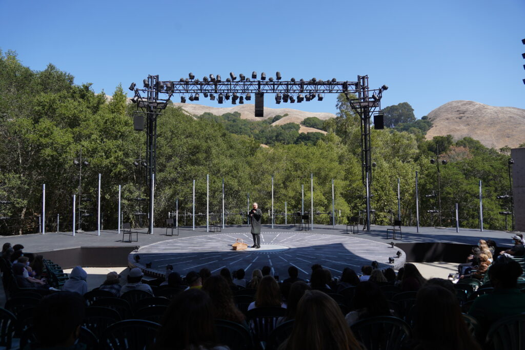 A person stands before an audience in the center of a circular outdoor stage, with mountains and trees in the background.