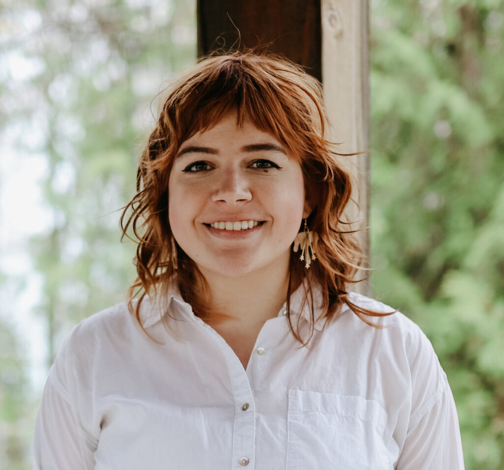 A smiling person of light skin tone with medium length red hair, wearing cloud and star earrings and a white button down shirt.
