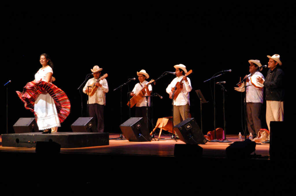 Chuchumbé, a band from Mexico who interprets and recreates music in verse and dance from the son jarocho tradition, performs on stage. Members stand in a line and play guitar, tambourine, and other instruments while singing, and one member dances in the front.