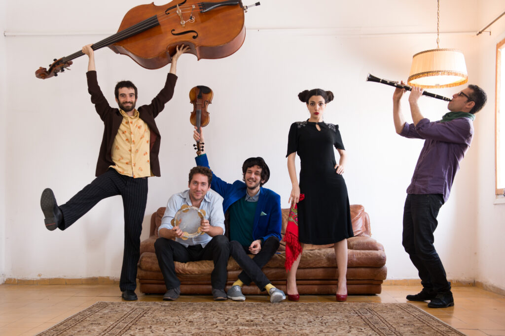 Baladino, a world music ensemble from Israel, pose in unique and humourous ways with their instruments. One is holding a standing bass above their head, one is holding a tambourine, one is holding a violin above the tambourine player's head, the singer poses with a scarf in hand, and one pretends to play a clarinet.