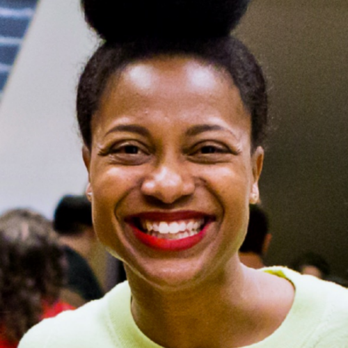 Headshot of a smiling person of medium dark skin tone, with black hair in a bun on top of their head, and wearing a bright yellow shirt.