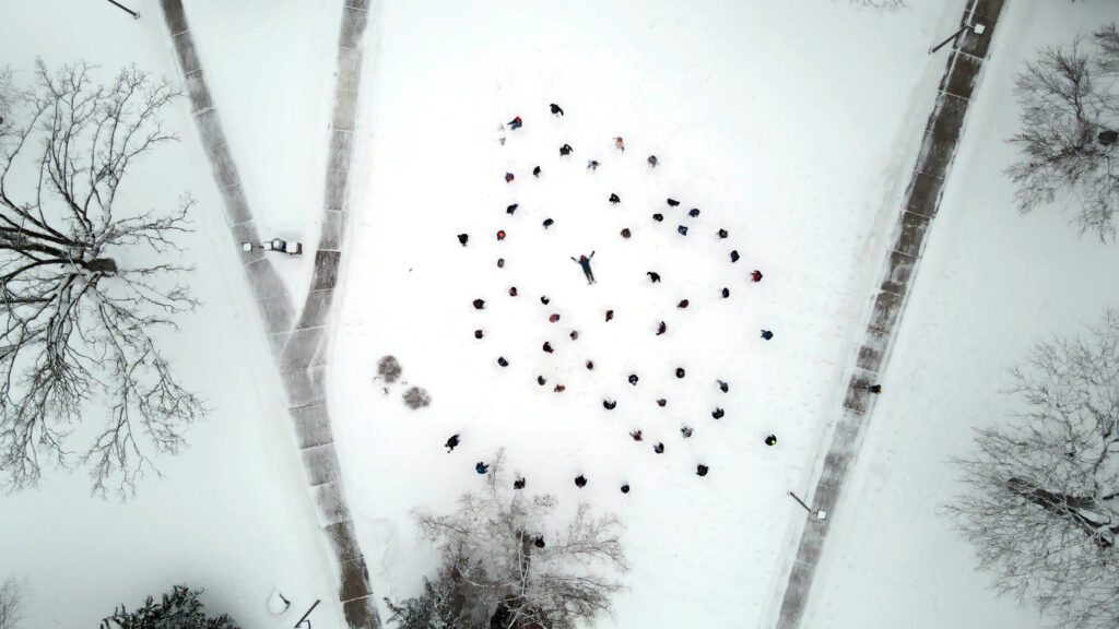 Bird's eye view of many people on snow-covered ground surrounded by sidewalk and trees. The people are standing and circling around on person in the center, who is laying down and sprawled out in snow angel fashion.