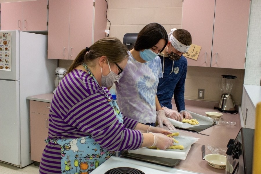 Three people in protective masks stand together in a kitchen, focused on braiding bread dough.