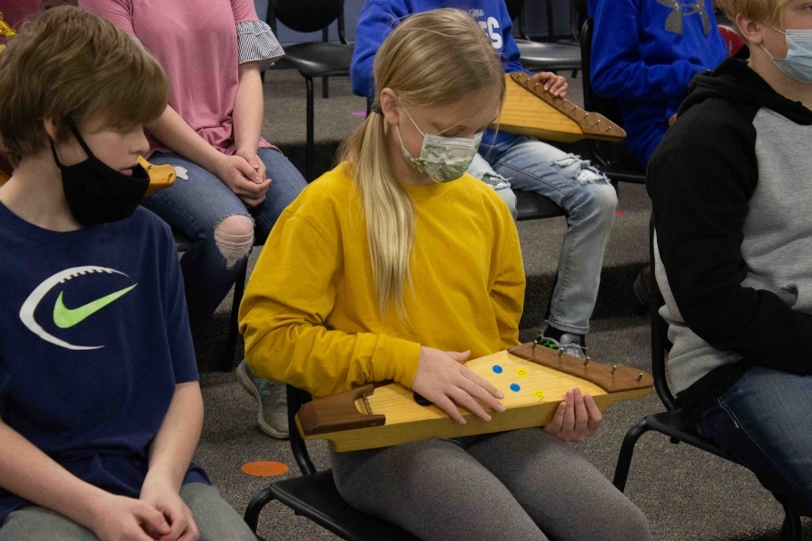A student in a surgical mask sits amongst other students, and is holding and observing a kantele, the Finnish national musical instrument.