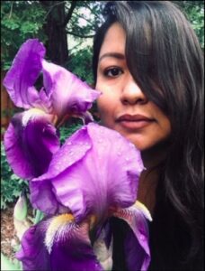Headshot of a softly smiling person of medium light skin tone, long black wavy hair covering part of their face, and posing next to a group of purple flowers.