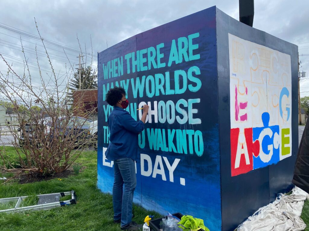 A person is adding paint to words on an outdoor mural that reads: "When there are many worlds you can choose...walk into each day." (The person is blocking part of the quote).