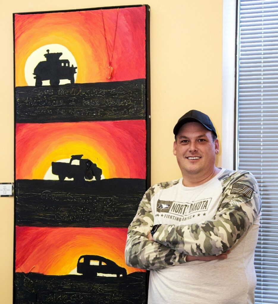 A smiling person of medium light skin tone, wearing a black baseball hat and a "North Dakota" shirt with camo sleeves, stands with arms crossed in front of a collection of three paintings. The paintings depict a silhouette of an army tank or trunk with a sunset behind it.