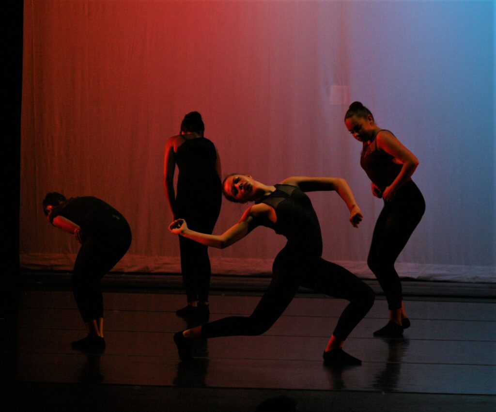 Four people dressed in black, dancing in front of a white backdrop that is lit by red and blue lights.