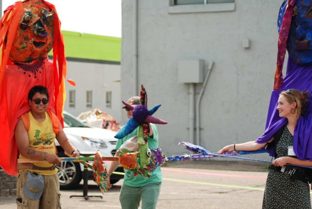 People perform with puppets made by artist Candace Ogborn in the parking lot at Interact Center for the Visual and Performing Arts.