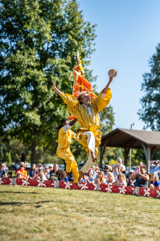 Two people in yellow outfits performing outside in front of children and adults. They leap joyously through the air with smiles on their faces.