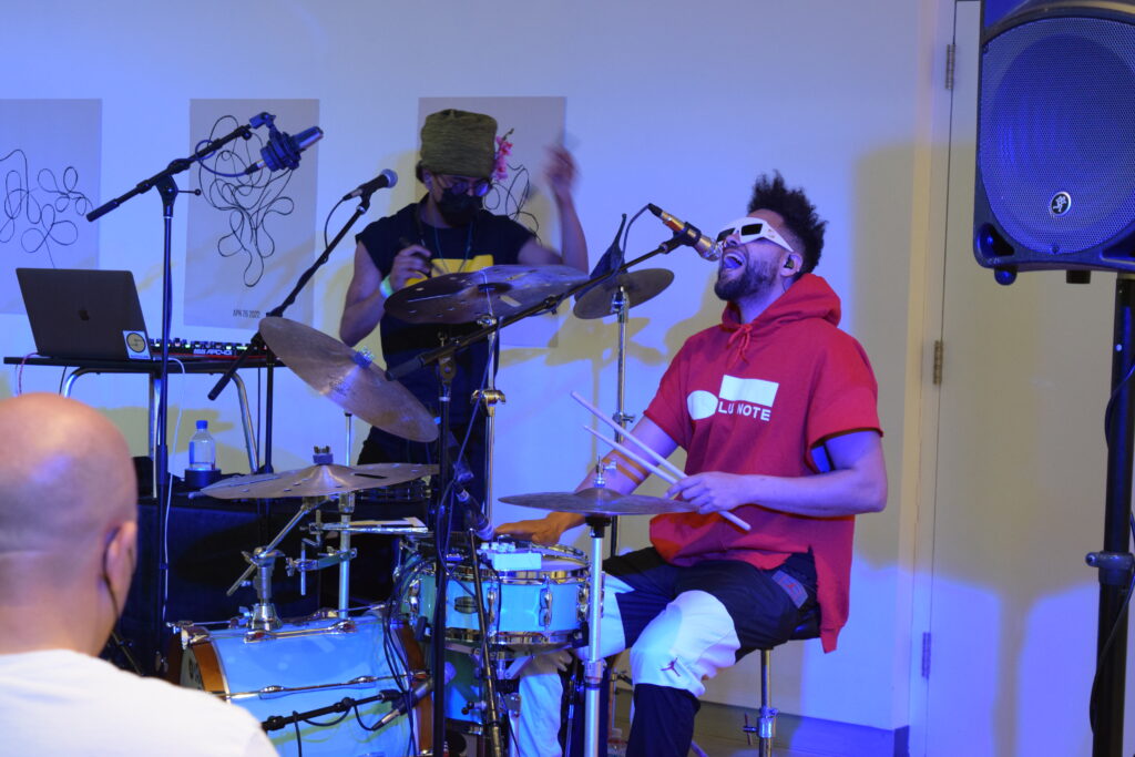 Two people performing; one is wearing white 3-D glasses and singing into a microphone, and the other is wearing sunglasses and a black protective mask and playing drums.