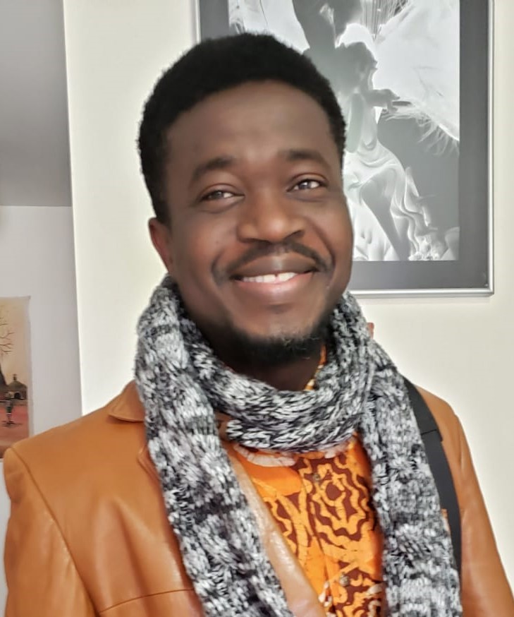 Headshot of a smiling person with dark skin tone and short black hair, mustache and beard, wearing a wool scarf over an orange jacket.