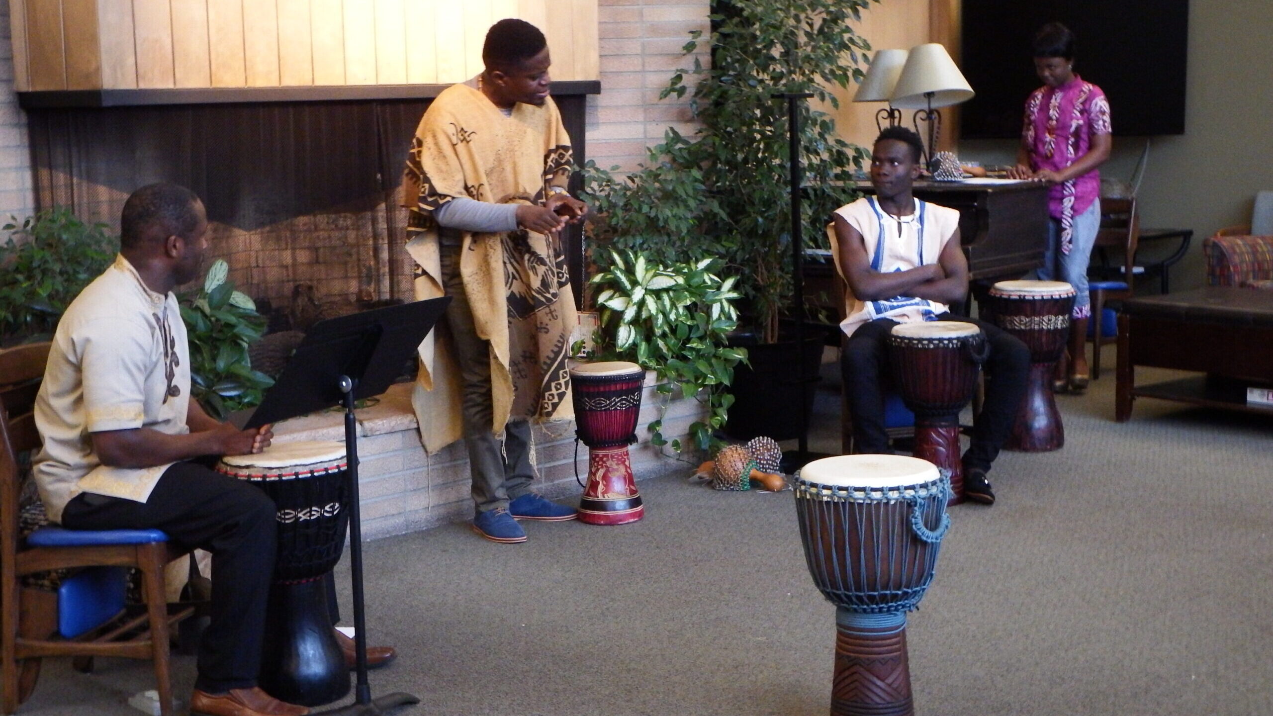 Three people performing with Djembe drums in front of a fire place.