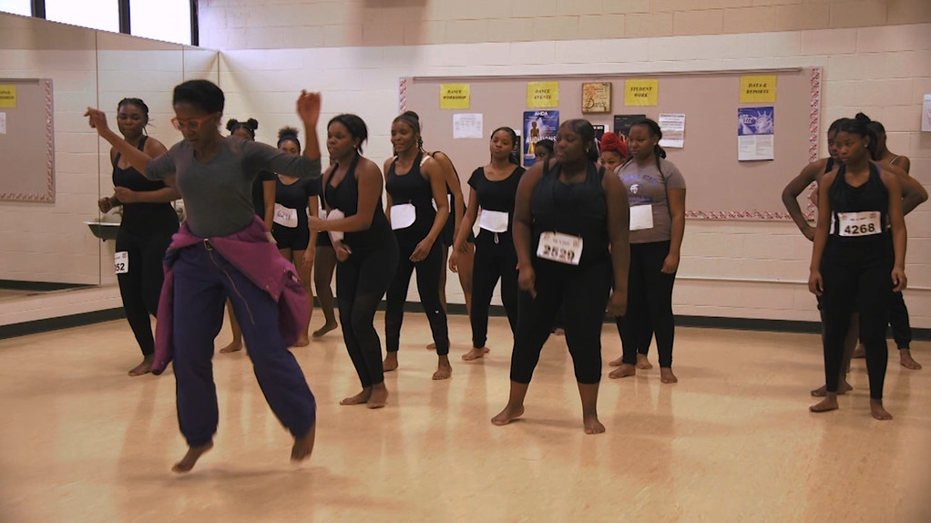 Many young people stand together in a dance studio, with audition numbers taped to their shirt. They're watching a teacher demonstrate a dance move at the front.