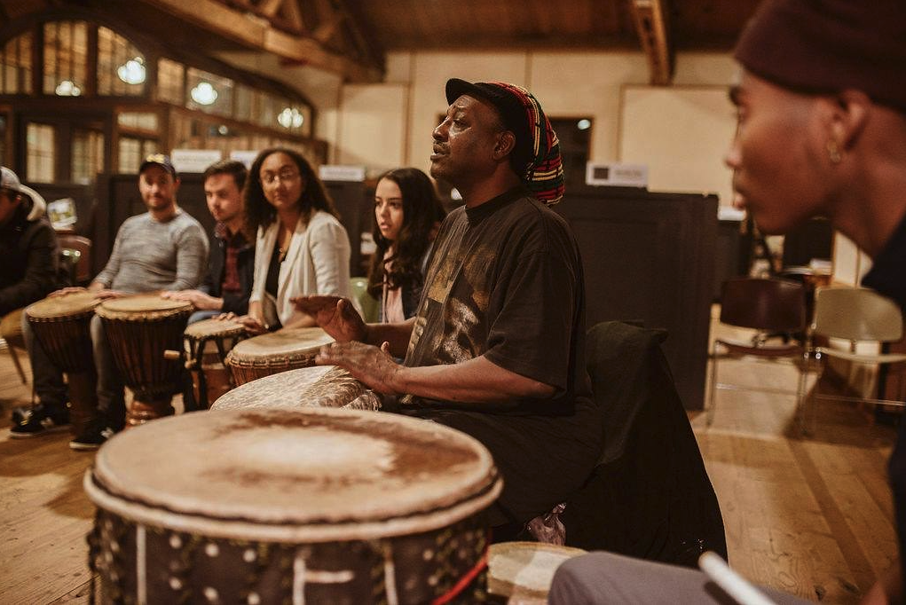 People sit together in a drum circle; one person plays while the rest watch.