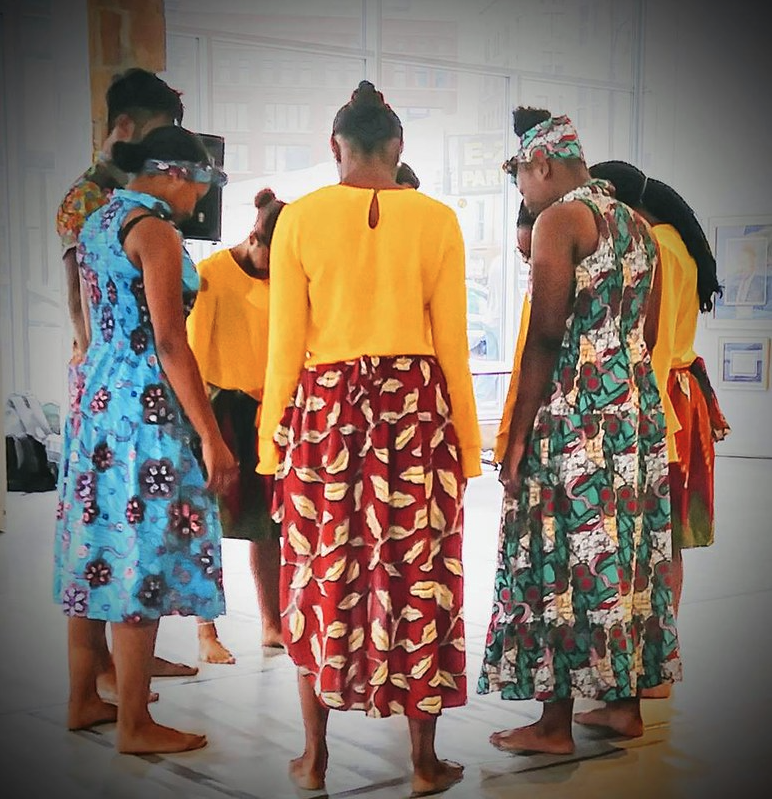 Heritage Work dancers wearing vibrant patterned dresses and skirts stand in a circle with faces angled down.