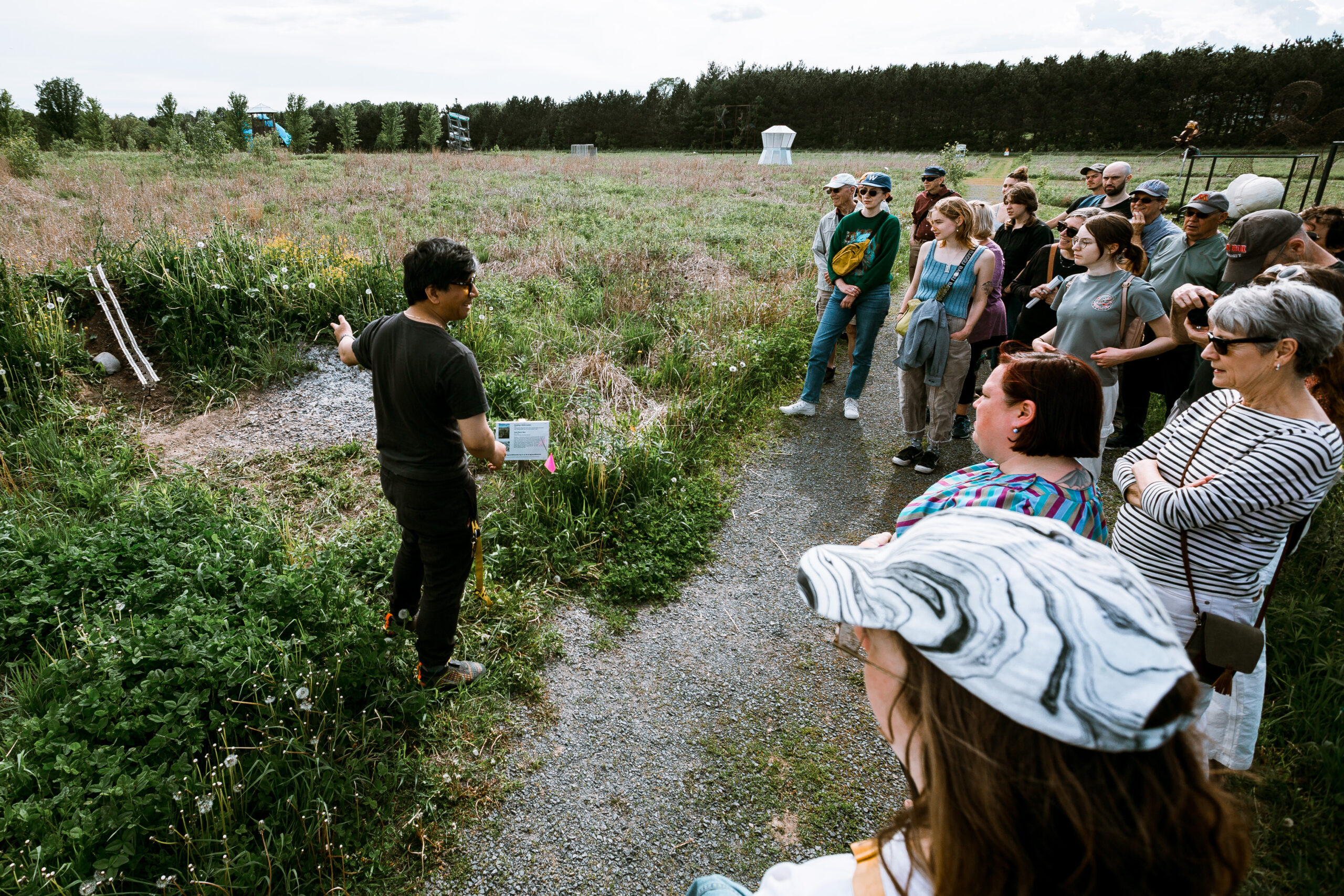 A person stands in front of a group of people outside, speaking and gesturing to an open area in the tall grass behind them.