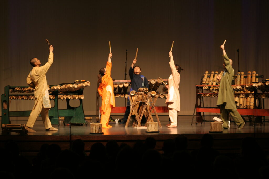 Bamboo Orchestra, a band from Japan, performs on stage, surrounded by bamboo instruments and holding bamboo sticks in the air.