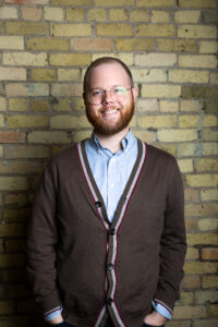 Headshot of a smiling person of light skin tone, short hair and a red beard, with round glasses and wearing a blue striped button down under a brown cardigan.