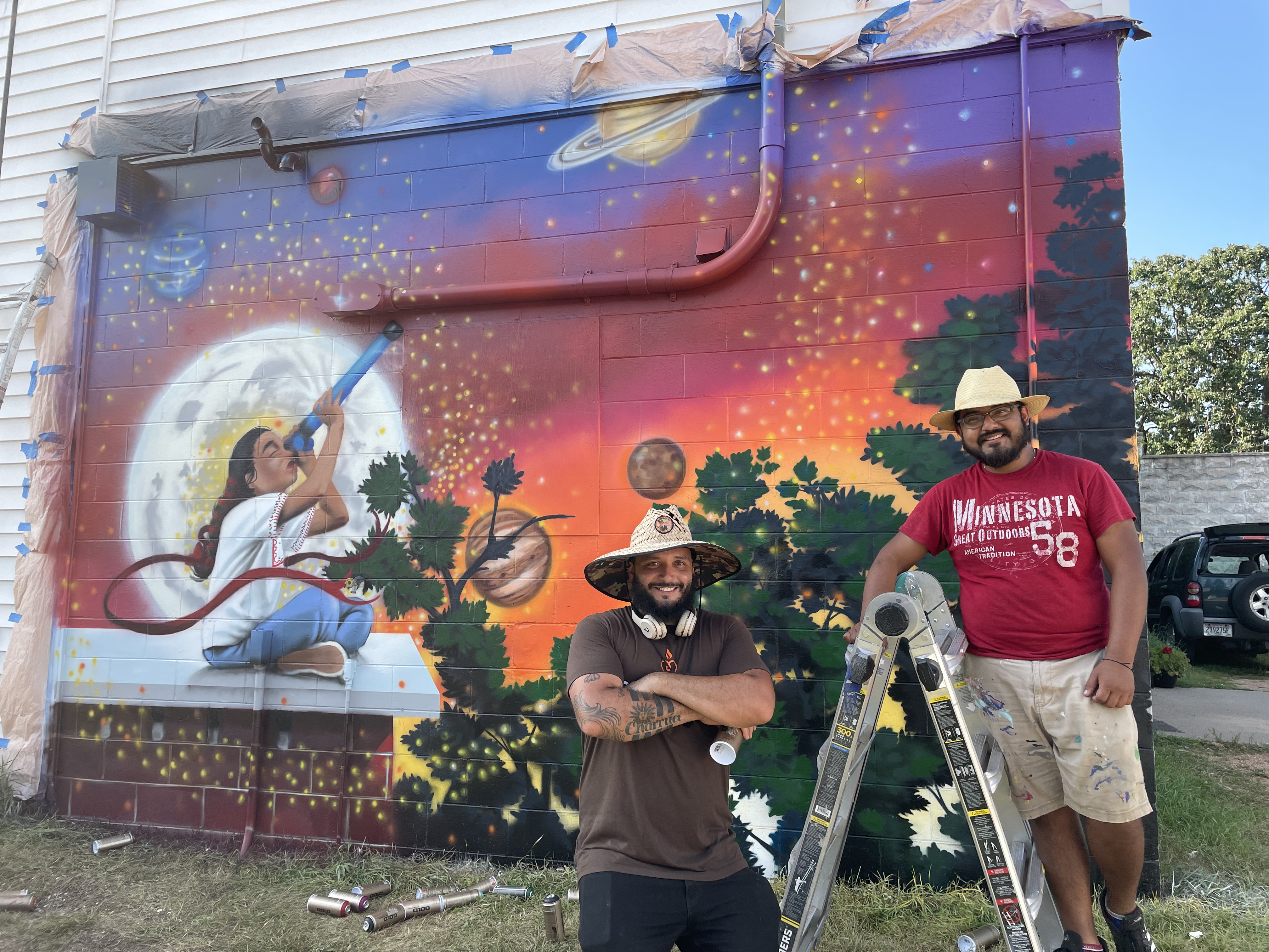 Two people wearing sunhats pose in front of a colorful mural on a brick wall. The mural depicts a young person sitting on a roof, holding a telescope up to a starry sky, with the moon big behind them and planets off in the distance.